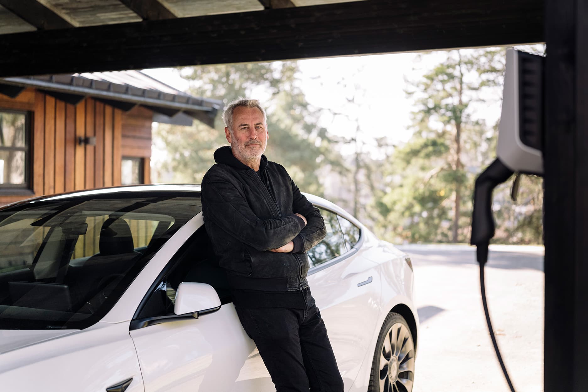 Anders Arnborger lutar sig mot sin Tesla 3, med en vit elbilsladdare på väggen