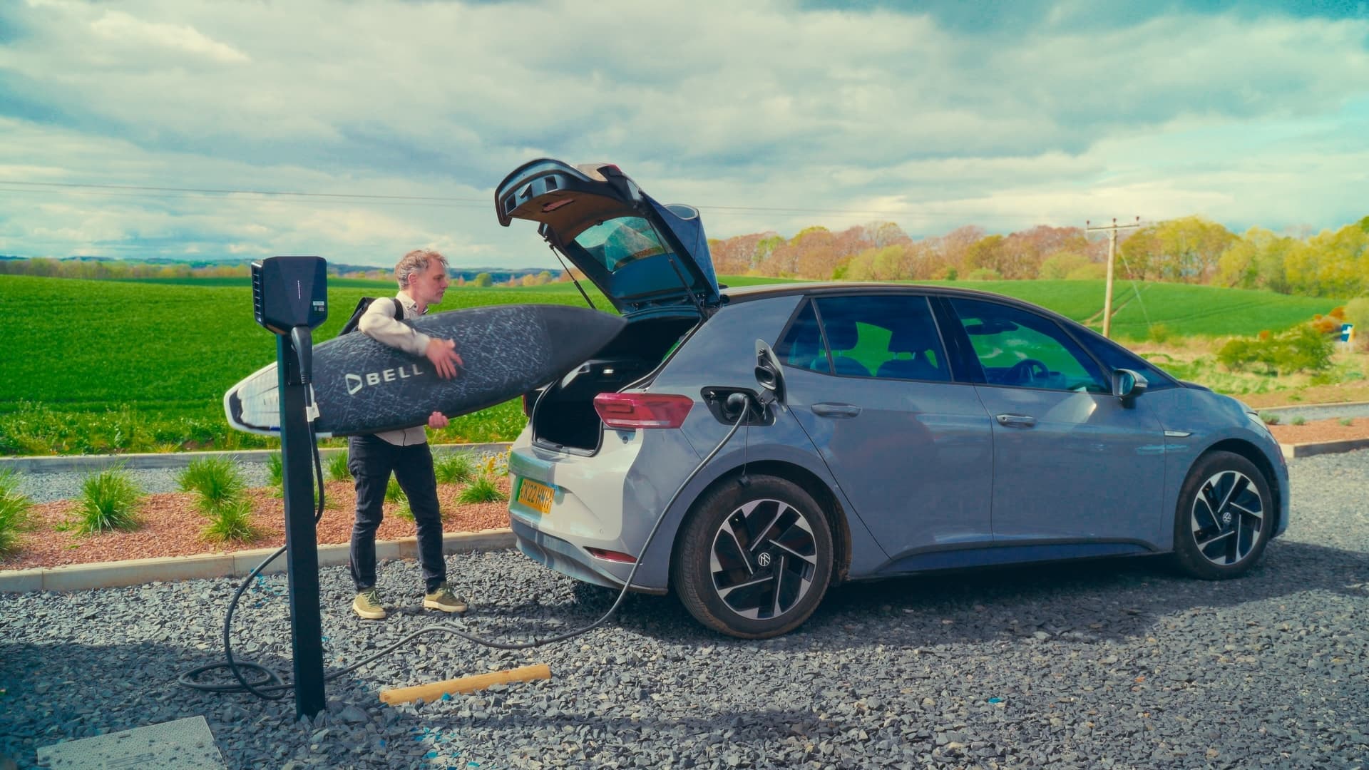 Man with surfboard beside black chargers on a base in a car park